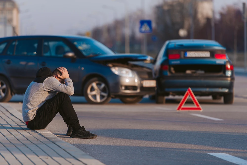 A young male seated on a curb with this head in his hands, with two vehicles involved in a t-bone accident in the background.