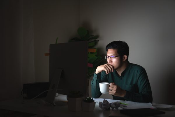 young man staring at a computer screen and holding a white coffee cup in a dark room