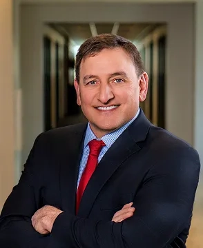 Smiling man in a dark suit and red tie, arms crossed in a hallway.