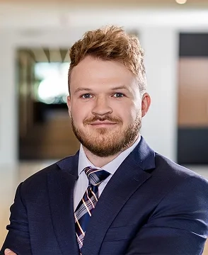 Professional headshot of a man with light brown hair and a beard, wearing a navy suit and striped tie, standing in an office setting with a neutral expression.