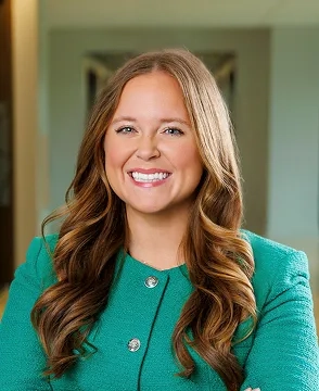 Professional head-and-shoulders portrait of a smiling woman with long wavy brown hair, wearing a teal blazer in a neutral indoor setting.