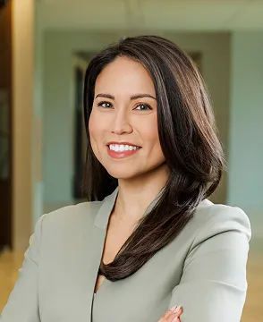 Professional woman with dark hair in a light grey blazer smiling confidently in an office setting.