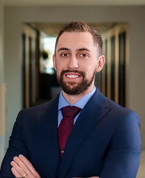 Professional headshot of a smiling man in a navy suit and burgundy tie, standing in a hallway with arms crossed.