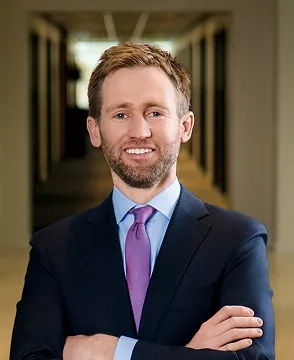 Professional headshot of a smiling man in a navy suit with a purple tie, arms crossed in a hallway.”,