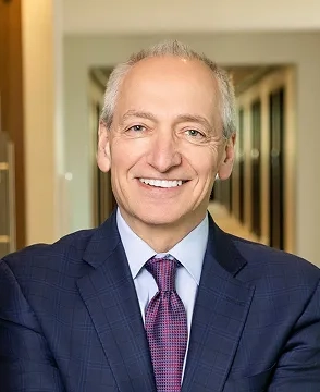 Smiling middle-aged man in a navy suit and pink tie, standing in an office hallway.