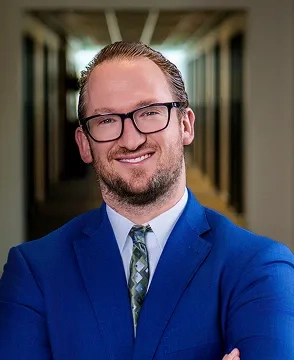 Professional headshot of a smiling man wearing glasses and a blue suit in a hallway setting with arms crossed in front of him