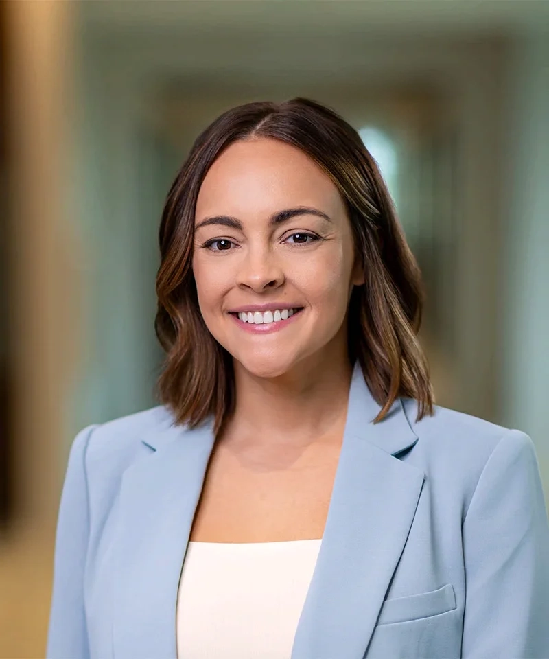 Professional headshot of a smiling woman with shoulder-length brown hair wearing a light blue blazer in a blurred office hallway.