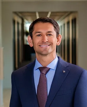 Professional head-and-shoulders photo of a smiling man in a navy suit and blue shirt in a blurred hallway backdrop.