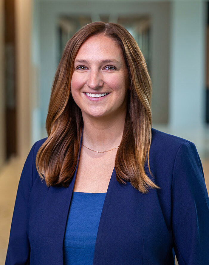 Smiling woman in a blue blazer and blue top, standing in a modern office.