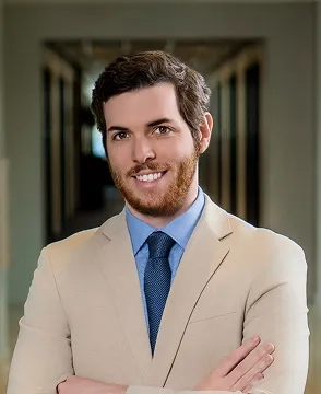 Man in a beige suit and blue tie smiles with arms crossed in a hallway corridor behind him.