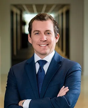 Smiling man in a navy suit and blue tie, arms crossed, standing in a hallway.