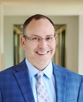 Smiling man in a blue suit, light blue shirt, pink striped tie and glasses, standing in a bright hallway.