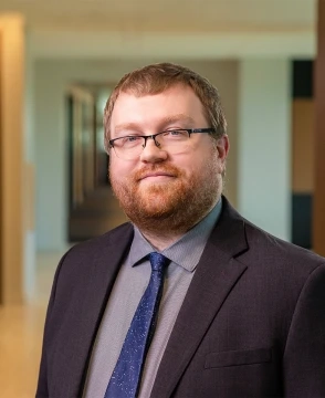 Portrait of a man with glasses and a beard in a dark suit and blue tie, standing in a bright hallway.
