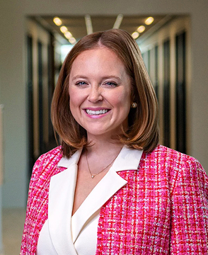 Professional headshot of a smiling woman with shoulder-length brown hair, wearing a pink tweed blazer and white blouse in a hallway.