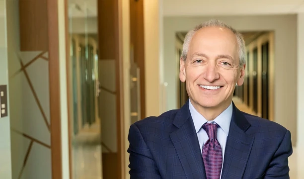 Smiling man in a navy suit and purple tie standing in a modern office hallway.