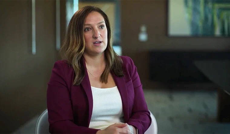 Woman in a burgundy blazer speaks during an interview in a modern office setting, hands folded in her lap.