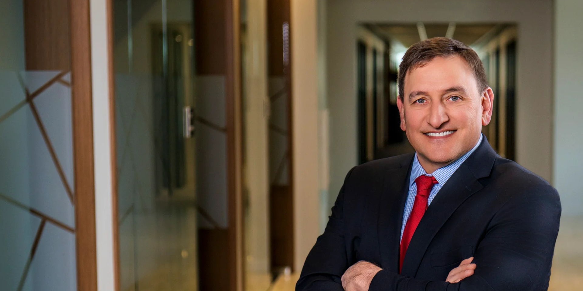 Smiling businessman in a navy suit and red tie, posed with arms crossed in a modern office hallway.