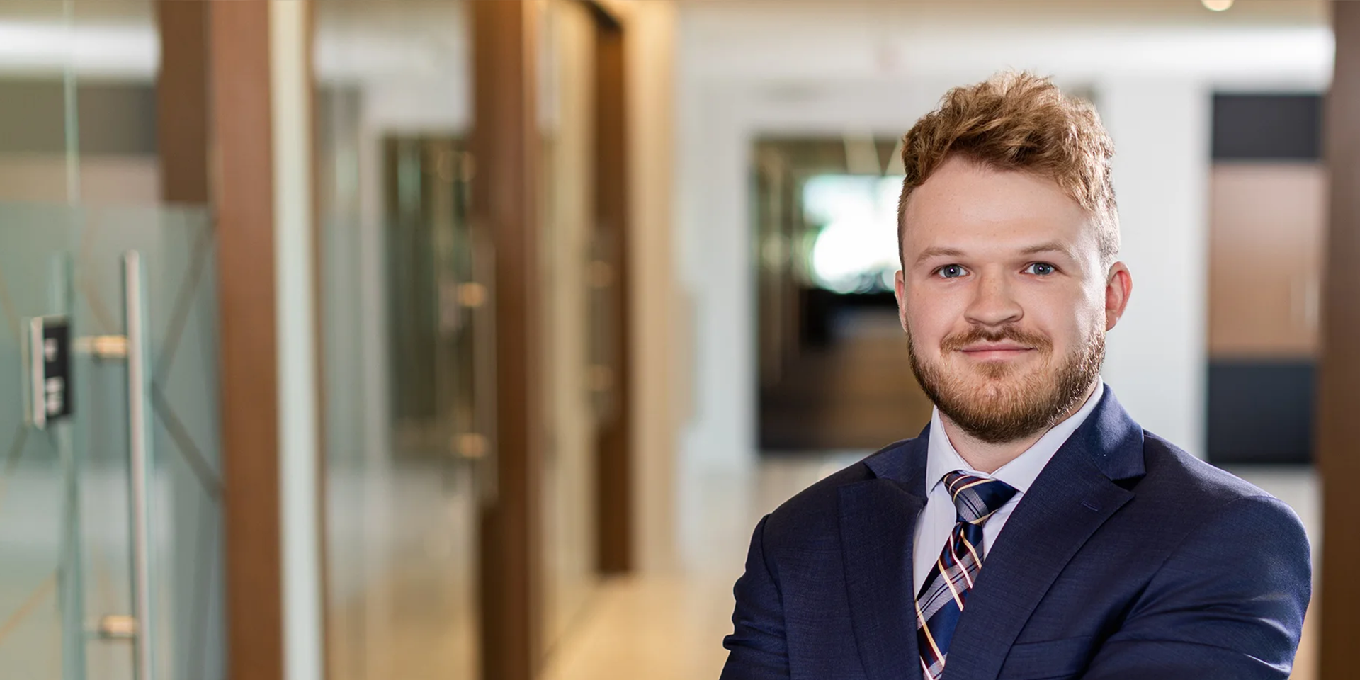 Portrait of a man in a navy suit smiling in a modern office hallway, posed for a professional headshot (office setting).