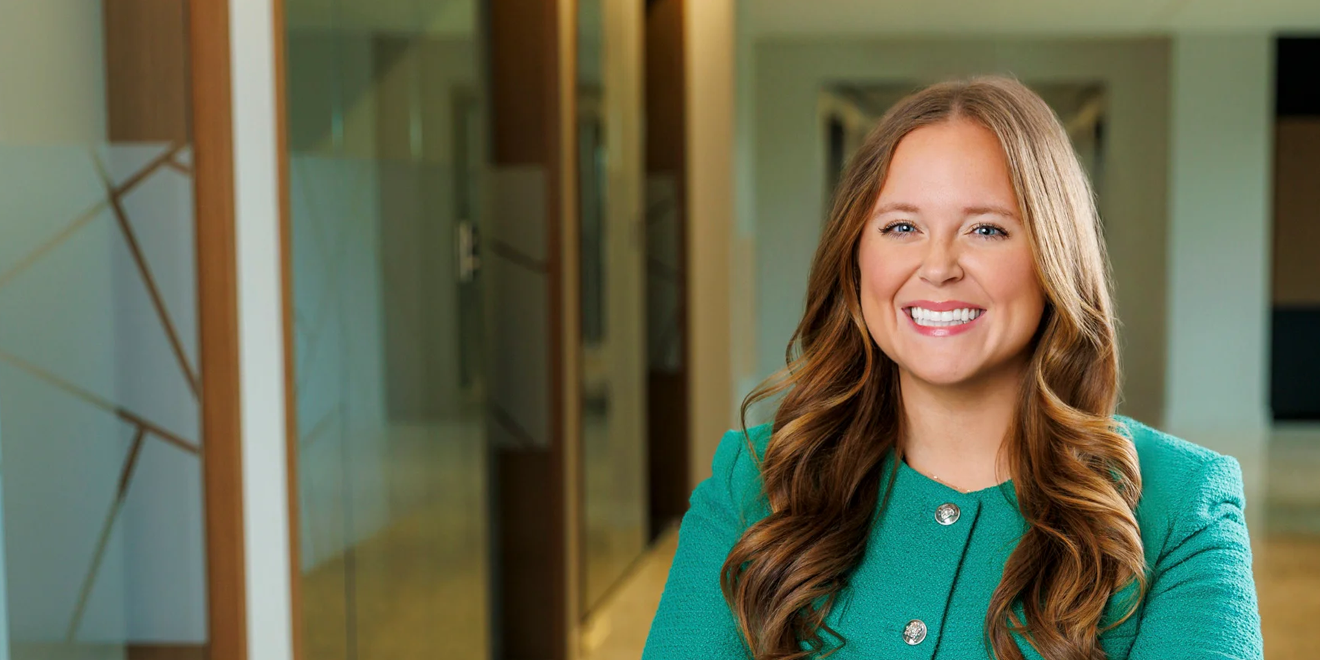 Smiling woman with long wavy hair in a teal blazer standing in a modern office hallway.