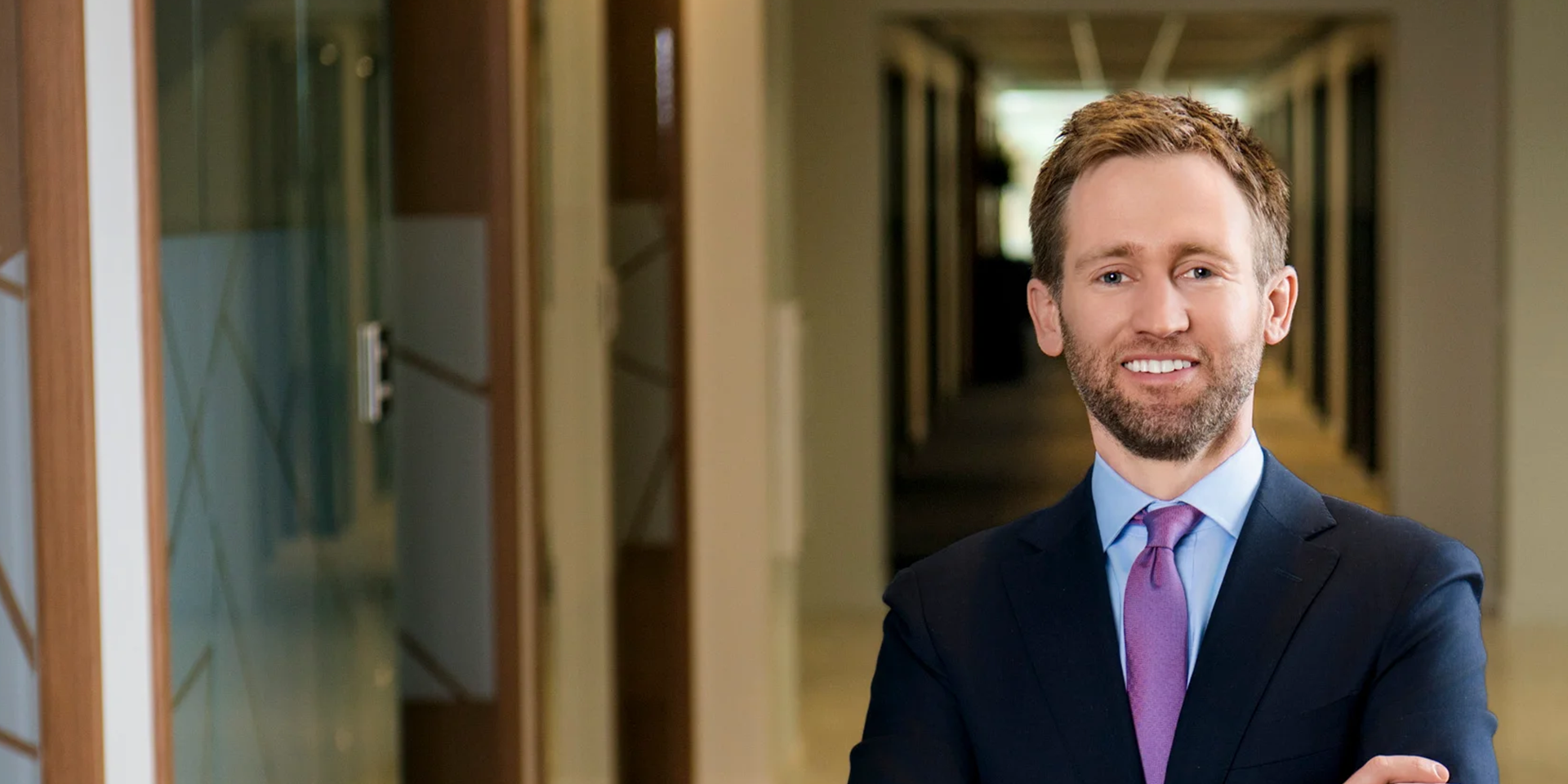 Smiling businessman in a navy suit and purple tie stands in a modern office hallway with glass walls on the left.