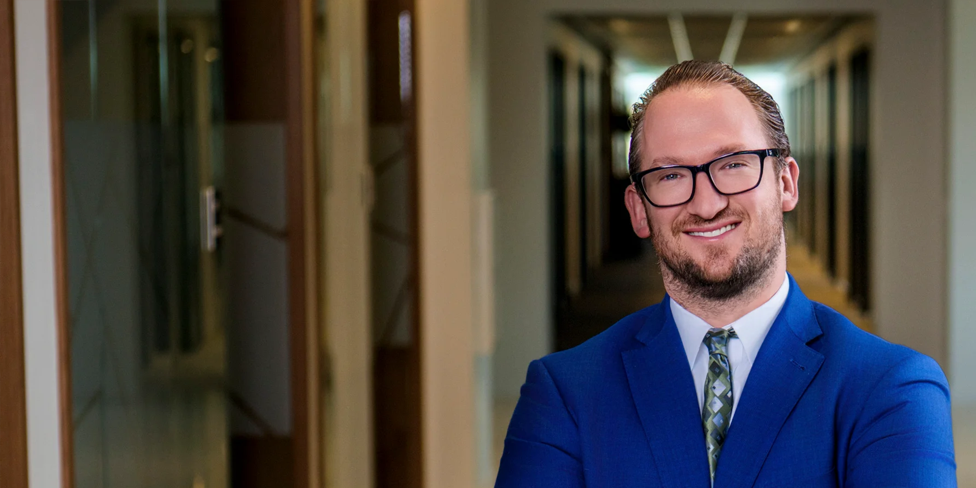 Smiling man in a blue suit and glasses stands in a modern office hallway, looking at the camera.