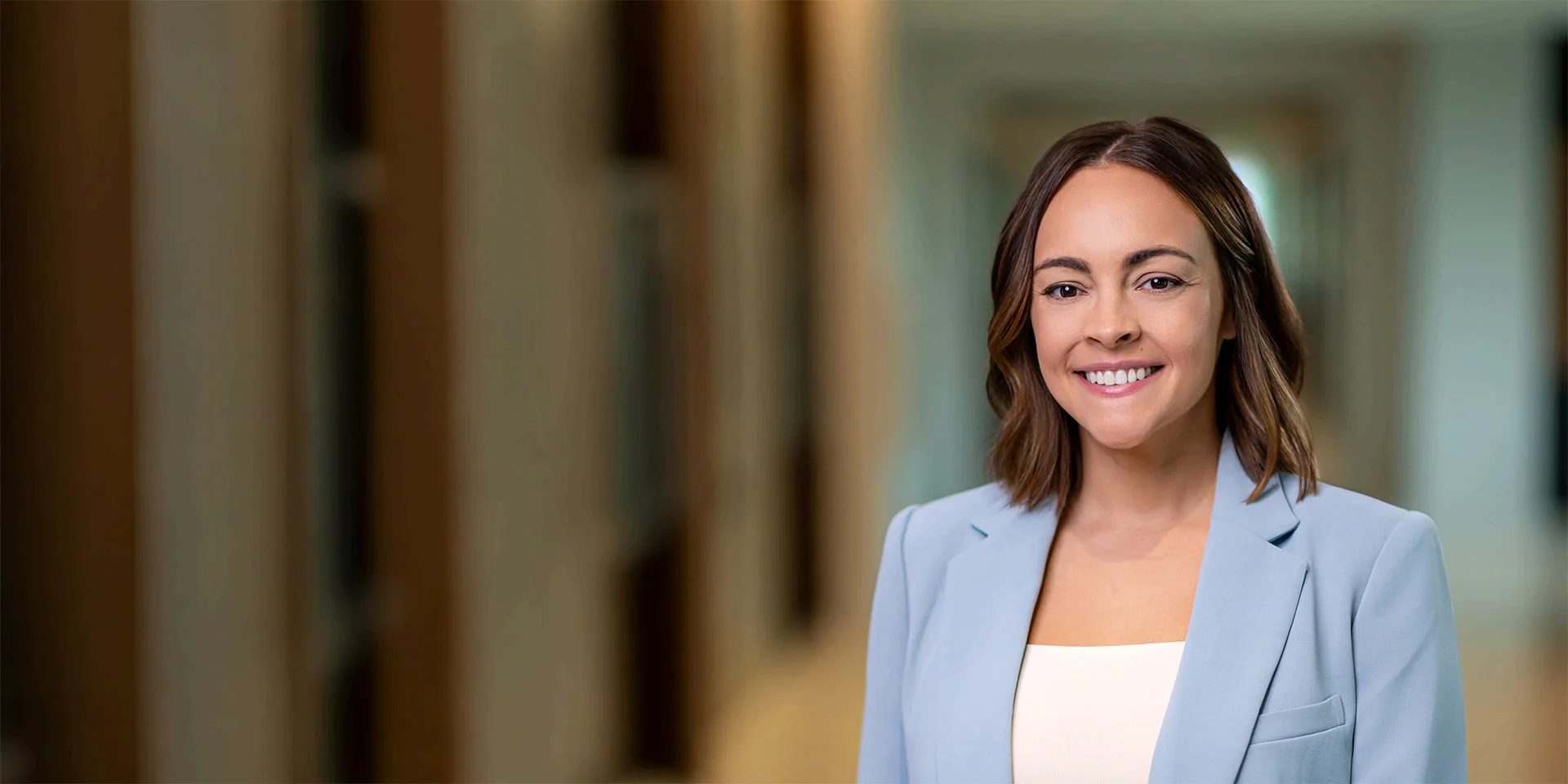 Professional woman in a light blue blazer smiles at camera in a corridor corridor setting as a headshot-style portrait.