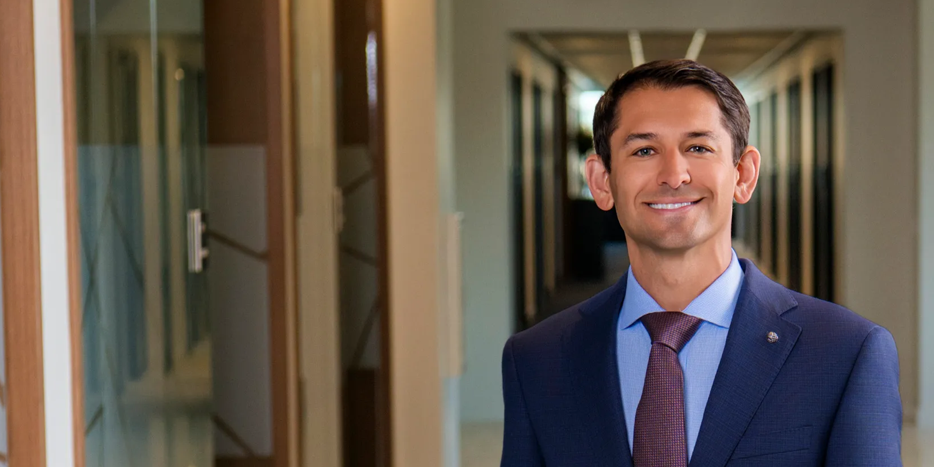 Professional portrait of a smiling man in a blue suit and maroon tie standing in an office hallway.