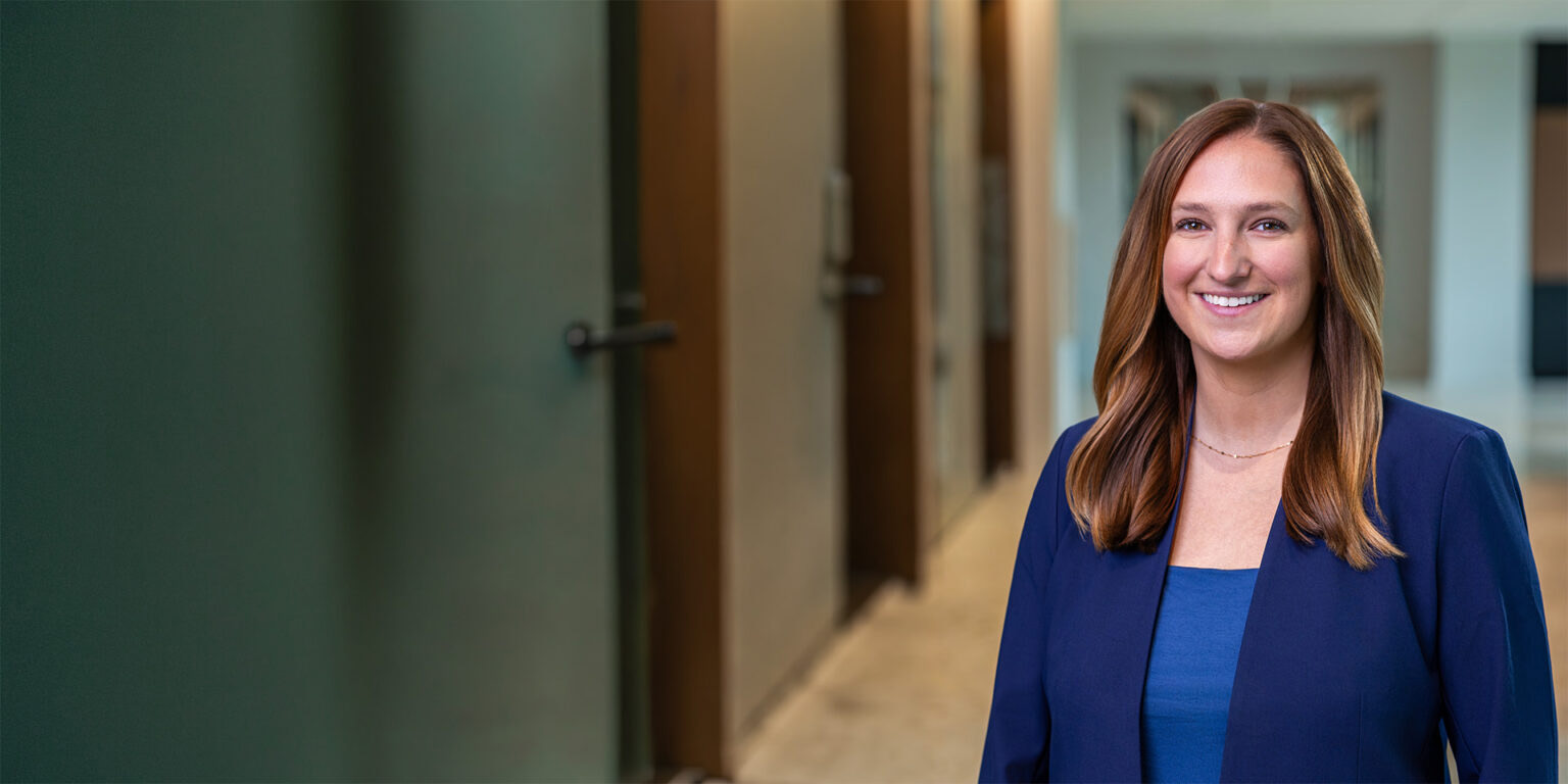 Smiling woman in a blue blazer standing in a modern hallway, conveying a professional portrait.