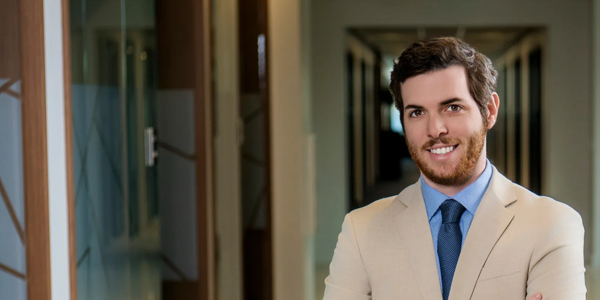 Smiling man in a beige suit posing in a modern office hallway.