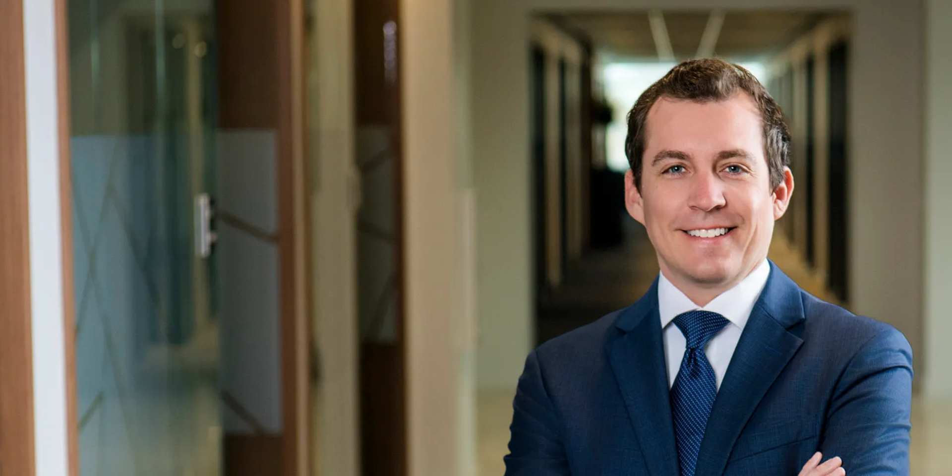 Smiling man in a blue suit standing with arms crossed in a office hallway.