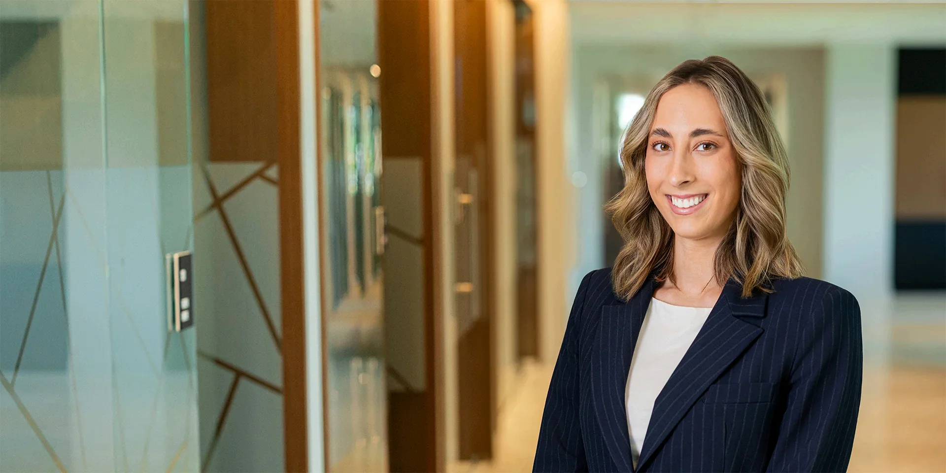 Smiling professional woman in a navy pinstripe blazer in a modern office hallway photographed as a portrait