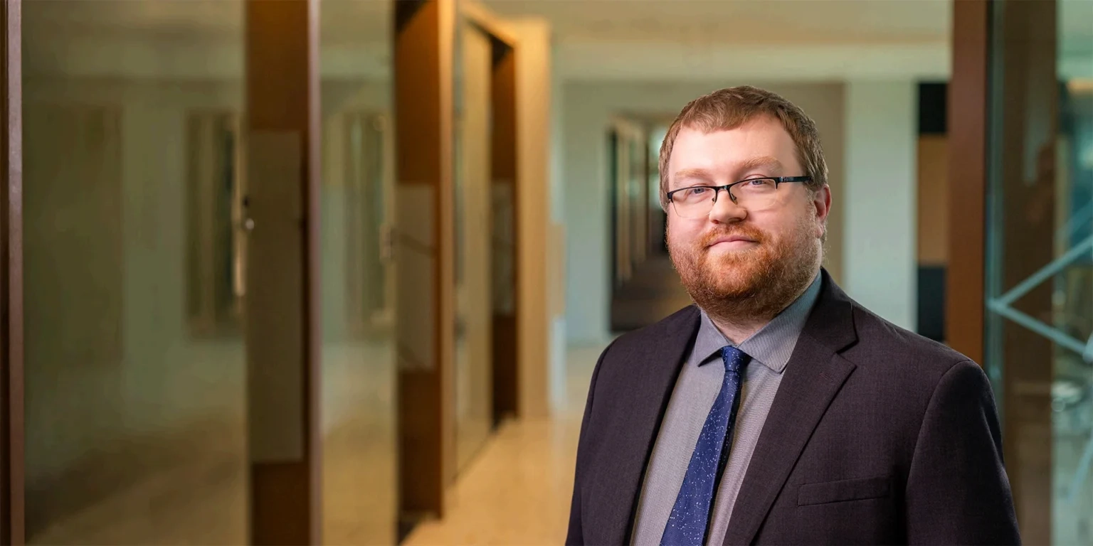 Businessman in a dark suit and blue tie standing in a modern office hallway.