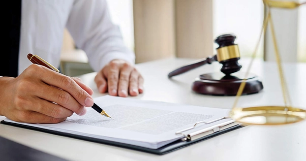 Man writing on a pad of paper with gavel and scale next to him on the desk.