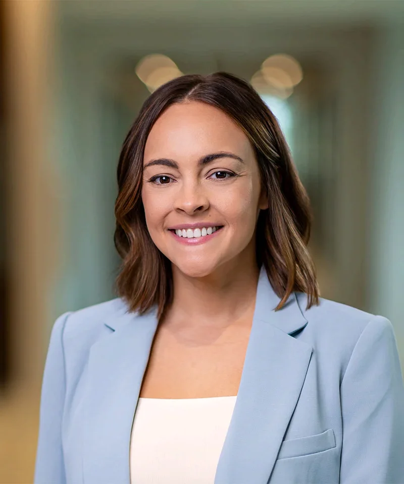 Professional headshot: smiling woman with shoulder-length brown hair in a light blue blazer, neutral office hallway background.