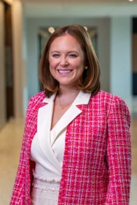 Smiling woman in a pink tweed blazer and white top standing in a bright hallway. A pearl earring is visible with a delicate necklace.