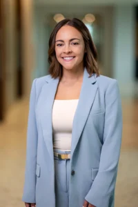 Professional woman wearing a light blue blazer and matching pants, smiling at the camera in a blurred hallway setting.