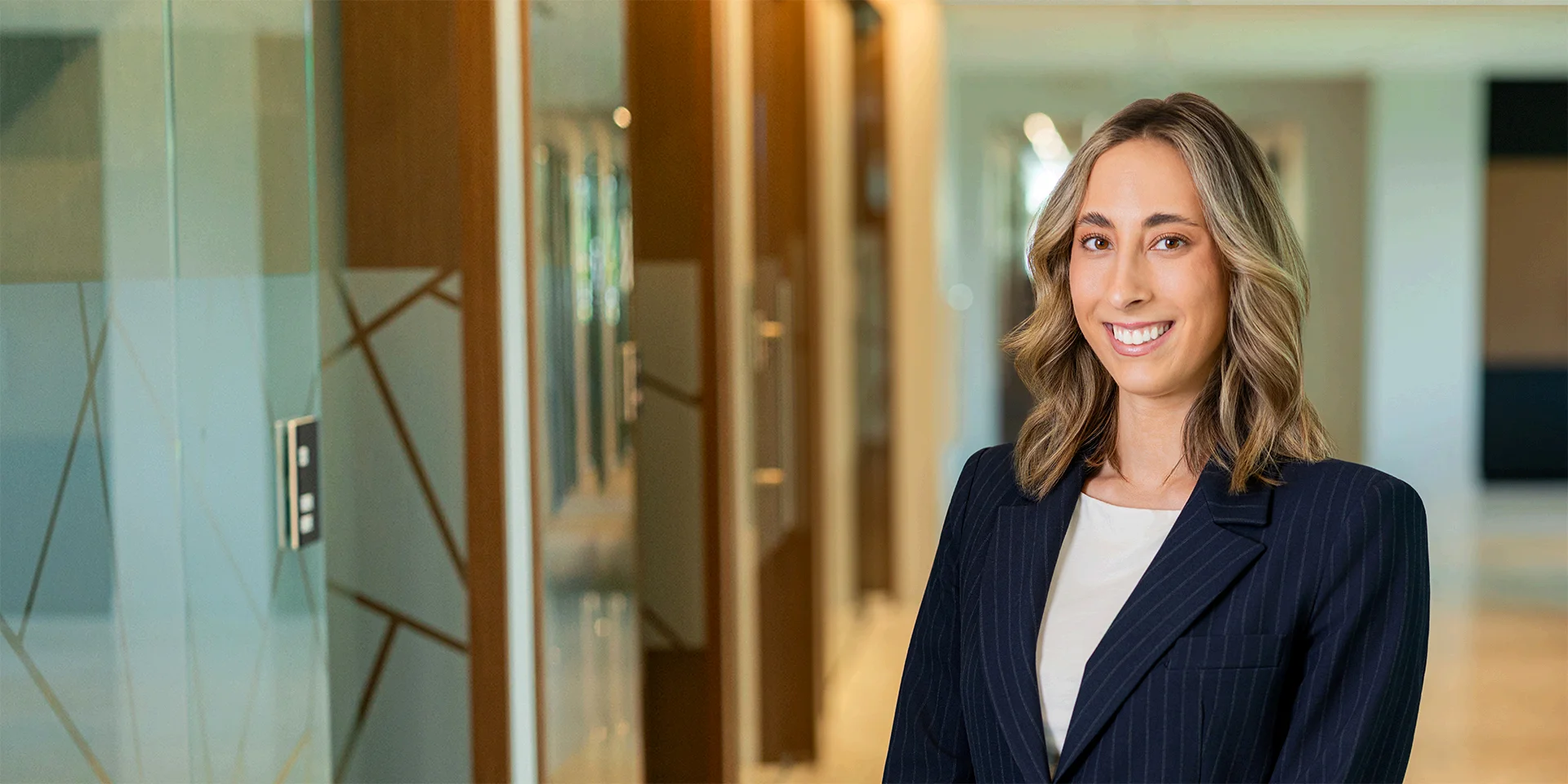 Professional woman in a navy pinstripe blazer smiles in a modern office hallway.