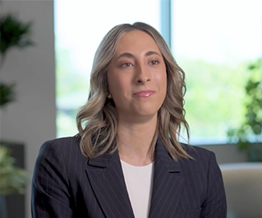 Professional woman in a pinstripe blazer seated in a bright office with greenery in the background, looking slightly to the side.
