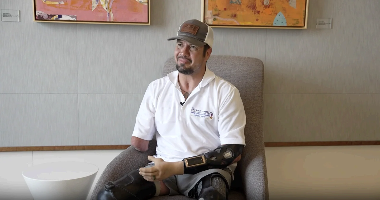 Man with a prosthetic arm and leg sits in a gray lounge chair, wearing a white polo and cap, microphone clipped to his shirt.