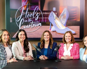 Five women sit at a panel table with microphones, smiling, in a conference room with a large screen showing 'Heels in the Courtroom' behind them.