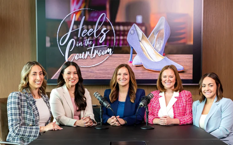 Five women sit at a panel table with microphones, smiling, in a conference room with a large screen showing 'Heels in the Courtroom' behind them.