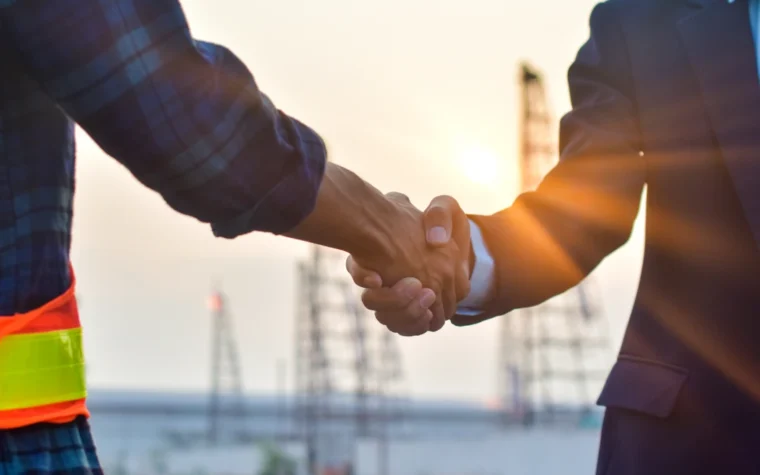 Handshake between a construction worker in plaid shirt with safety belt and a businessperson in a suit at a construction site during sunset, symbolizing a deal or partnership.