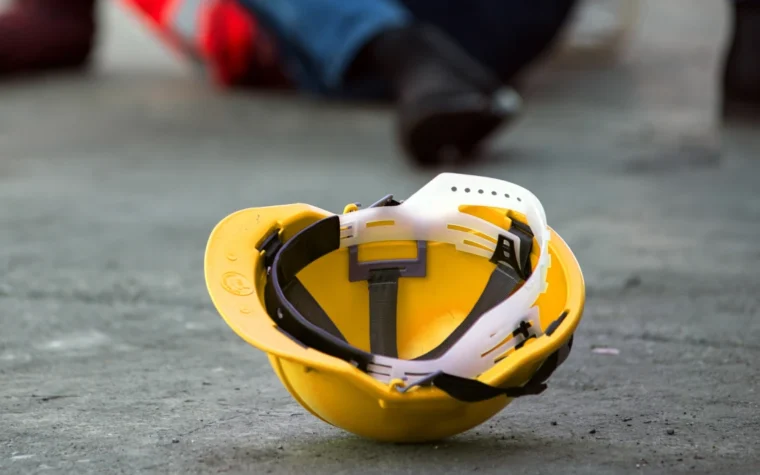 Yellow construction helmet with a white face shield resting on concrete, blurred worker in the background.
