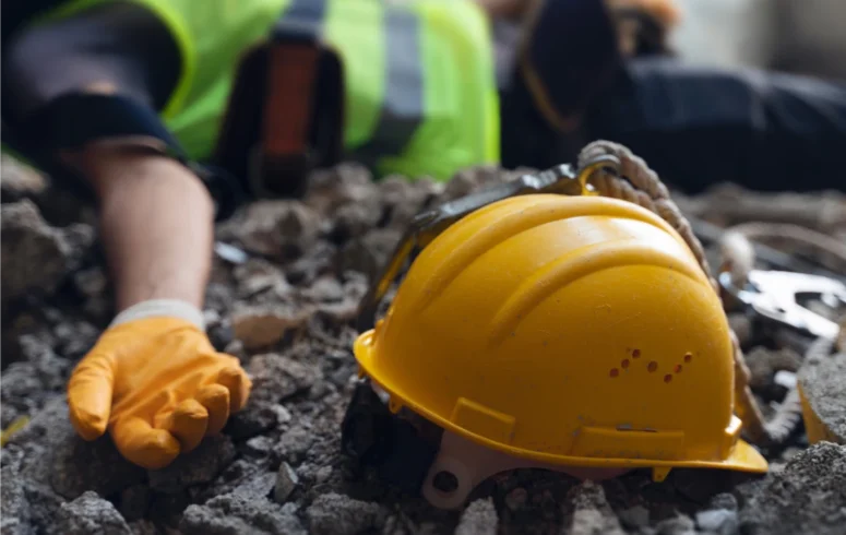 Yellow safety helmet resting on rubble with an orange gloved hand nearby at a construction site.