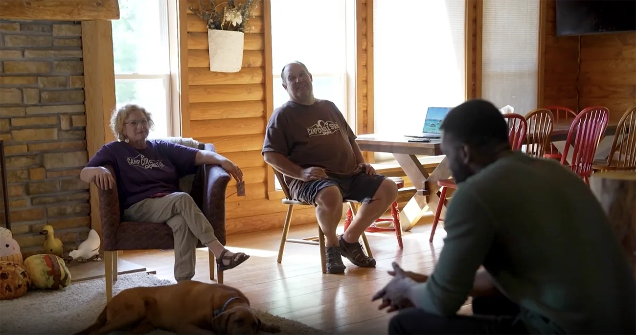 Two older adults sit in a cozy wooden cabin living area while a man in the foreground talks to them; a dog naps on the rug nearby.