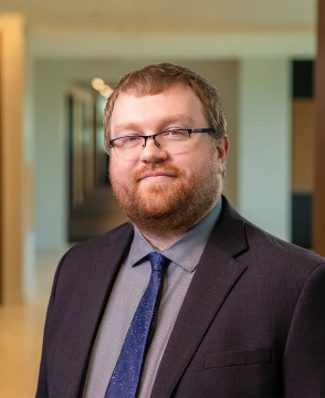 Professional head-and-shoulders portrait of a bearded man in a dark suit and blue tie, standing in a well-lit hallway with blurred background.