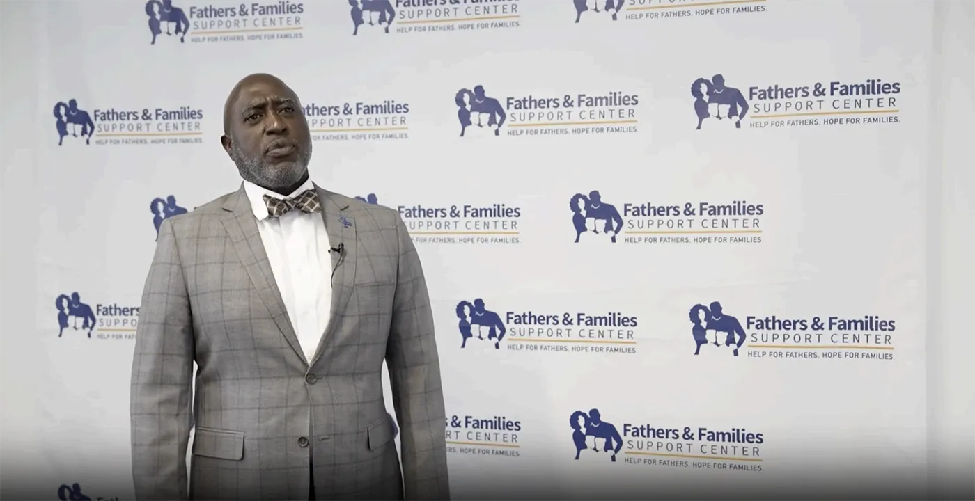 Man in a gray checkered suit and bow tie stands at a backdrop labeled Fathers & Families Support Center, speaking to an audience with a lapel mic attached.