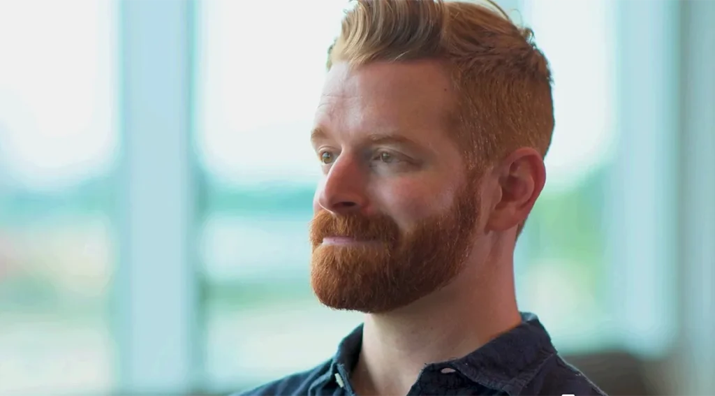 Portrait of a man with a red beard and short blond hair looking to the left in a bright, window-lit room.