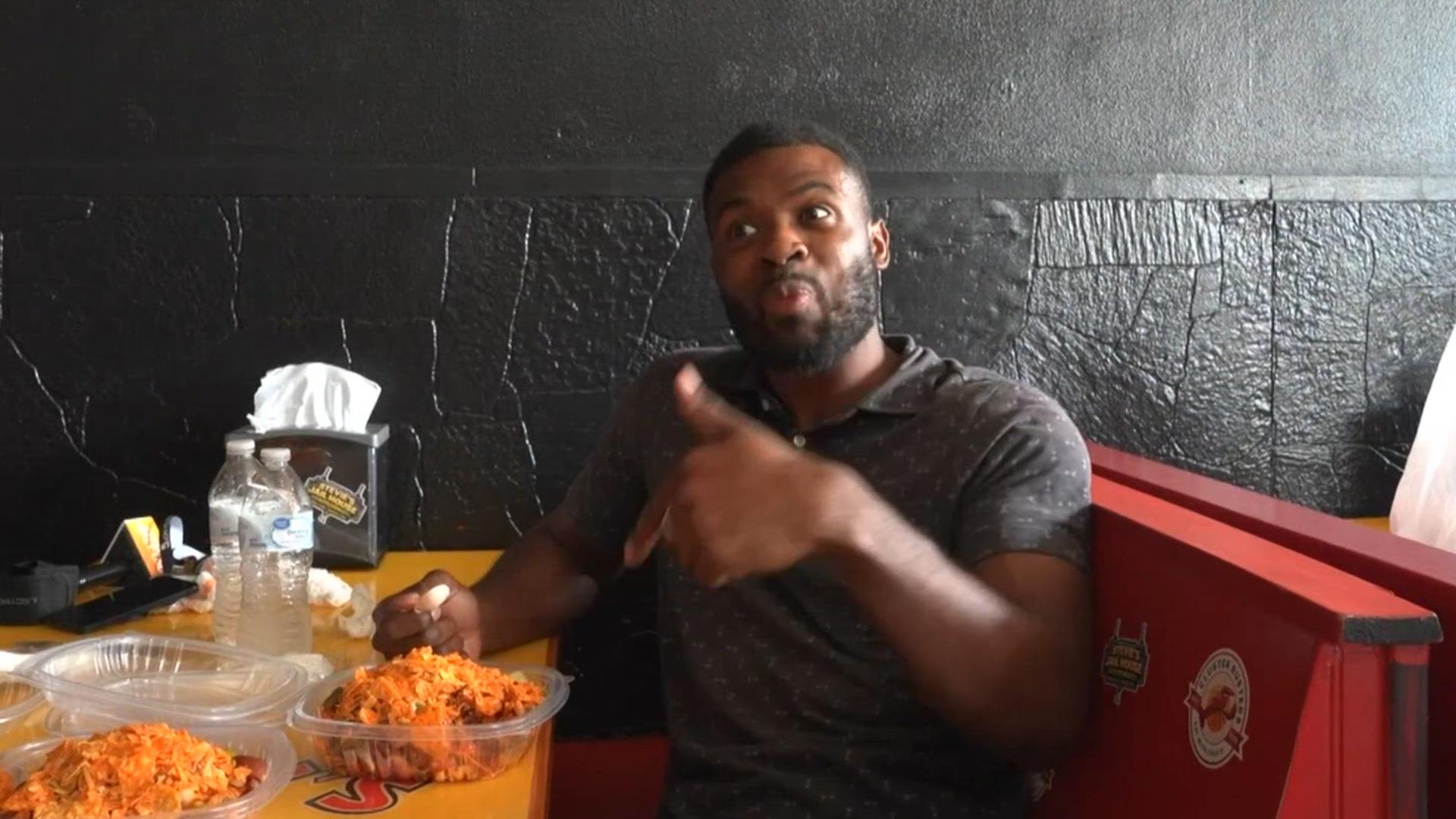 Man in a dark polo shirt sits at a table, gesturing with his hand as he talks, with bowls of chopped orange salad and drinks in front of him against a black textured wall.