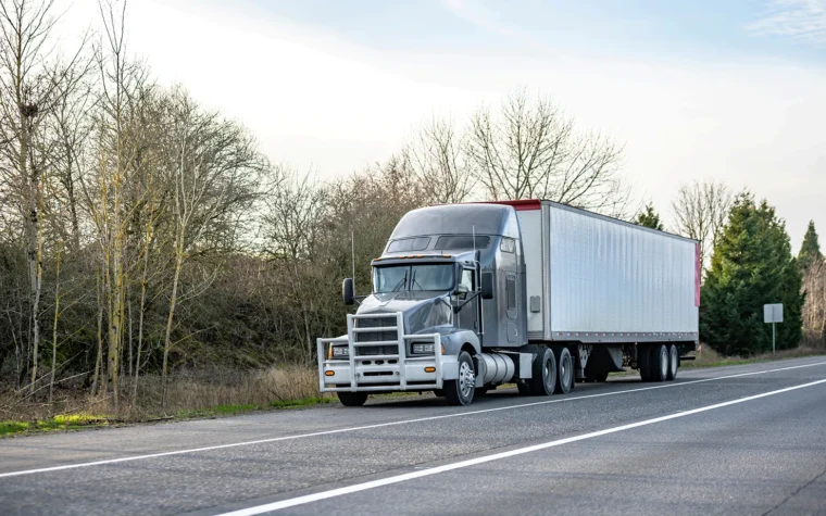 Gray semi-truck with a large silver trailer parked on the roadside beside a two-lane highway, leafless trees in the background.